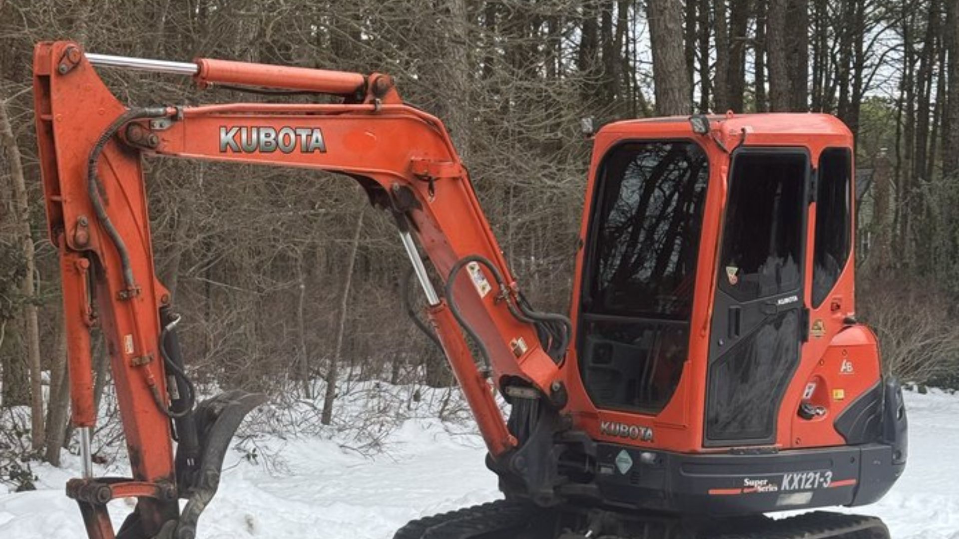 An orange Kubota excavator parked in a snowy, wooded area.