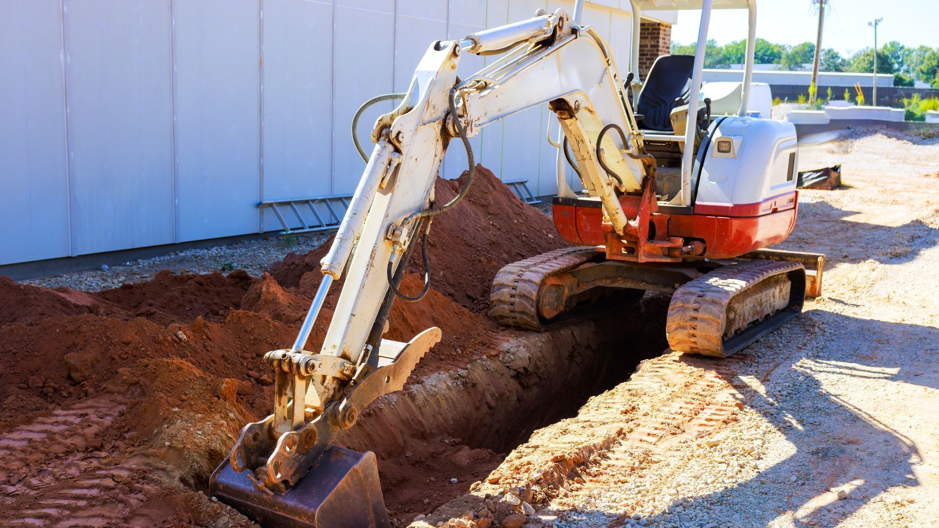 A white and orange mini excavator digs a trench in red dirt next to a light-colored building on a construction site.