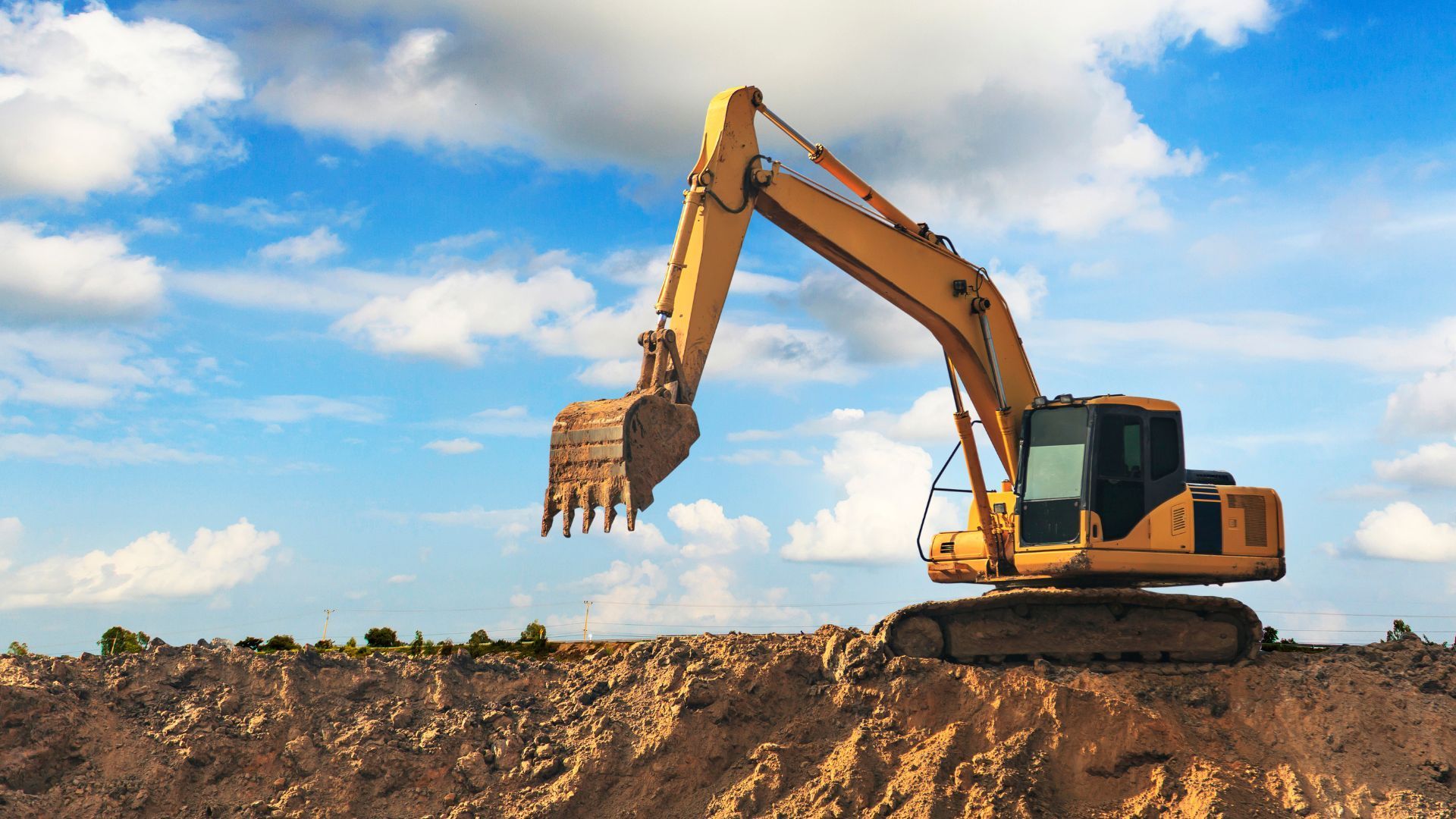 A yellow excavator sits atop a dirt pile against a blue sky with scattered clouds.