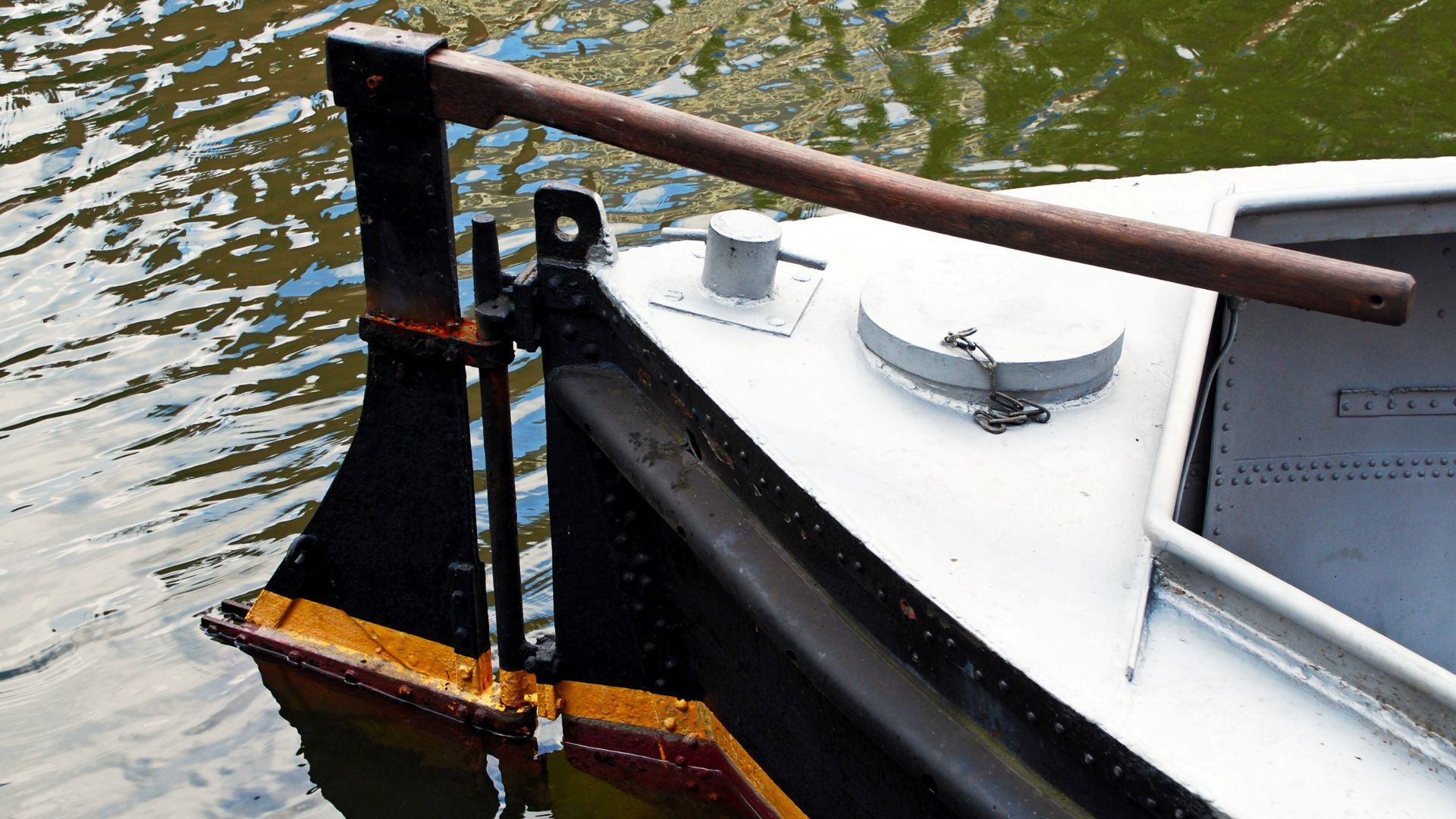 Close-up of a boat's rudder and tiller assembly attached to the stern above calm water.