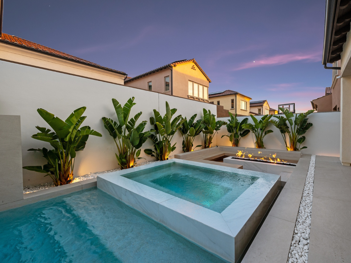 Backyard pool and spa area with tropical plants, a fire pit, and homes in the background under a sunset sky.