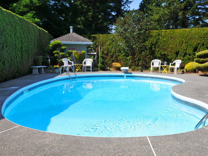Swimming pool surrounded by green hedges, patio, and chairs under a sunny sky.