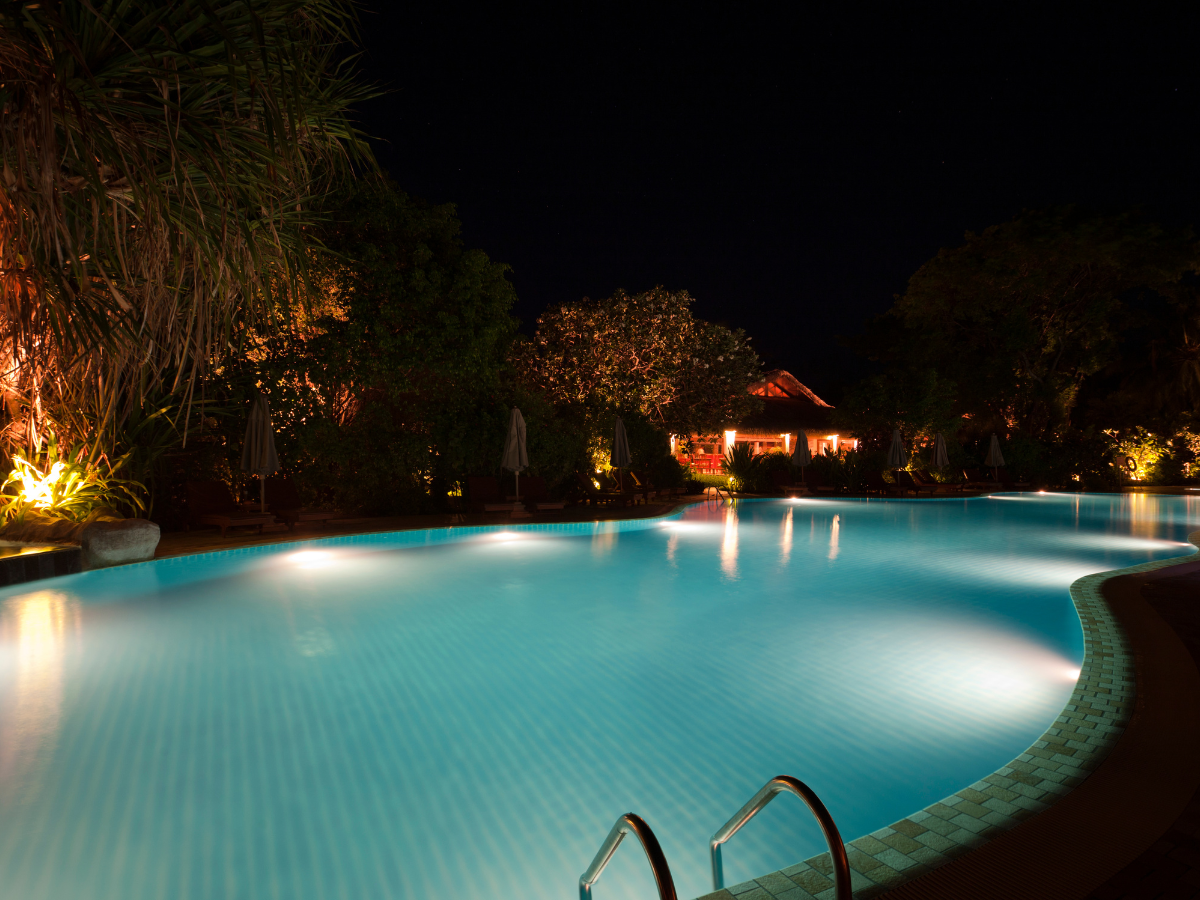 Nighttime pool with underwater lights, surrounded by trees and a building in the background.
