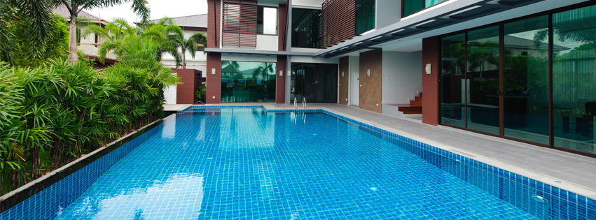 A long, rectangular swimming pool in front of a modern home with large windows and lush greenery.