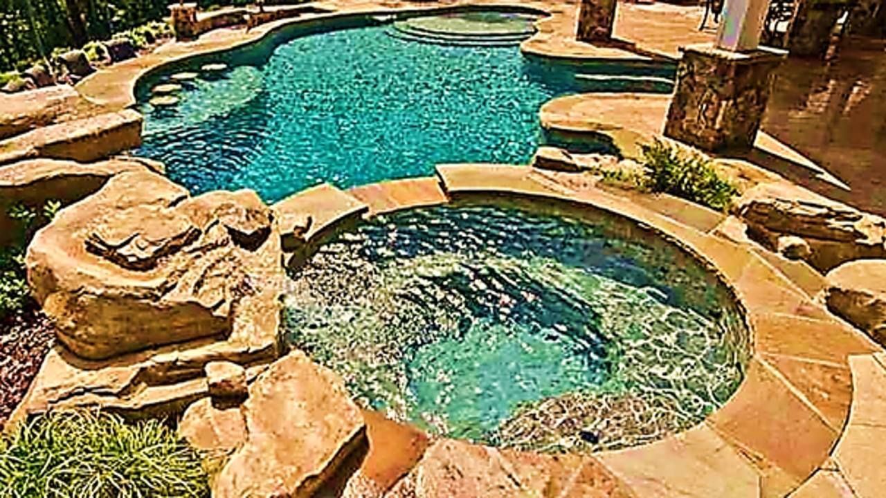 Pool and hot tub surrounded by stonework, with turquoise water and landscaping.