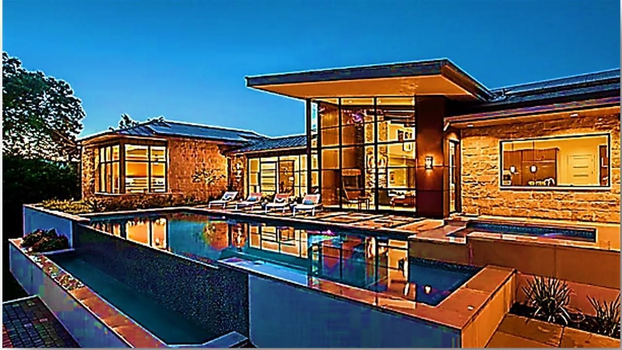Modern home with an infinity pool at dusk. Large windows, stone facade, and a flat roof.