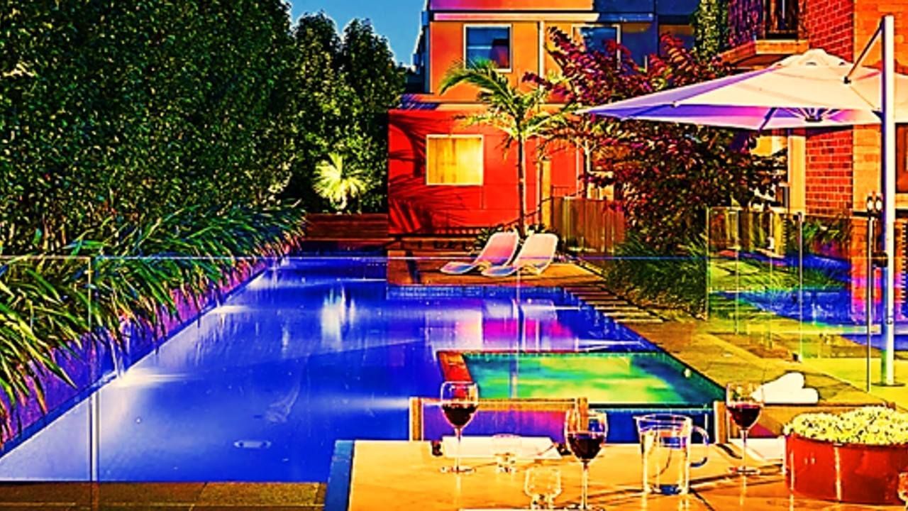 Pool lit with blue lights next to a table with wine glasses, an umbrella, and a colorful building.
