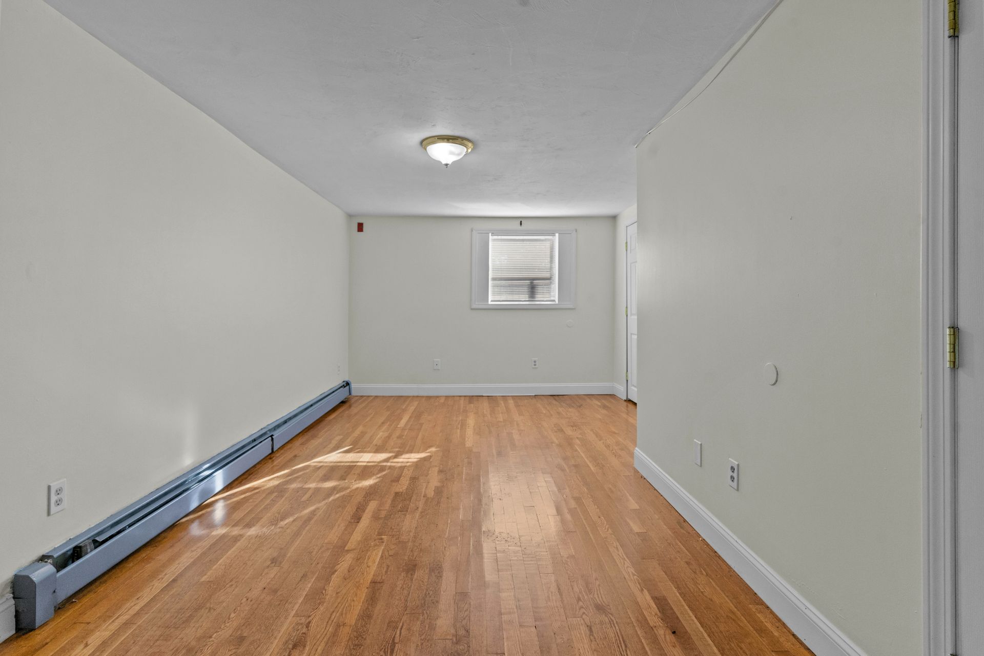 An empty room with hardwood floors, white walls and a window.