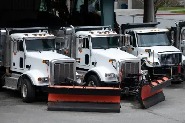 A row of snow plows are parked in a parking lot.