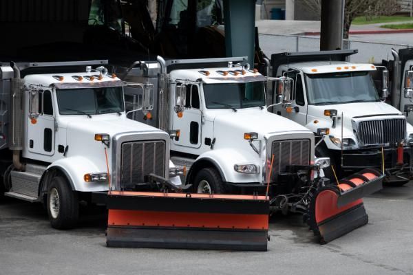 A row of snow plows are parked in a parking lot.