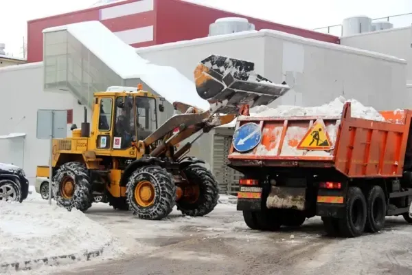 A yellow tractor is loading snow into a dump truck in a commercial parking lot.