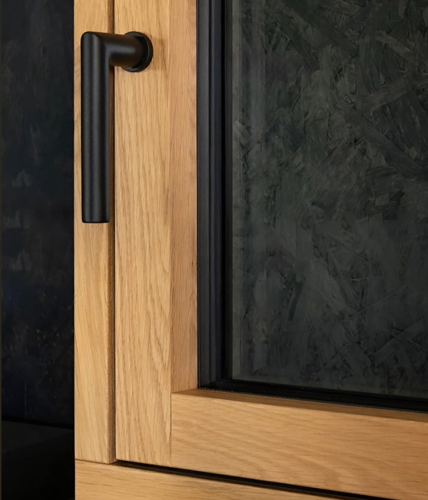 Primo piano di una maniglia nera opaca su un telaio di finestra in legno chiaro, su uno sfondo scuro e strutturato.