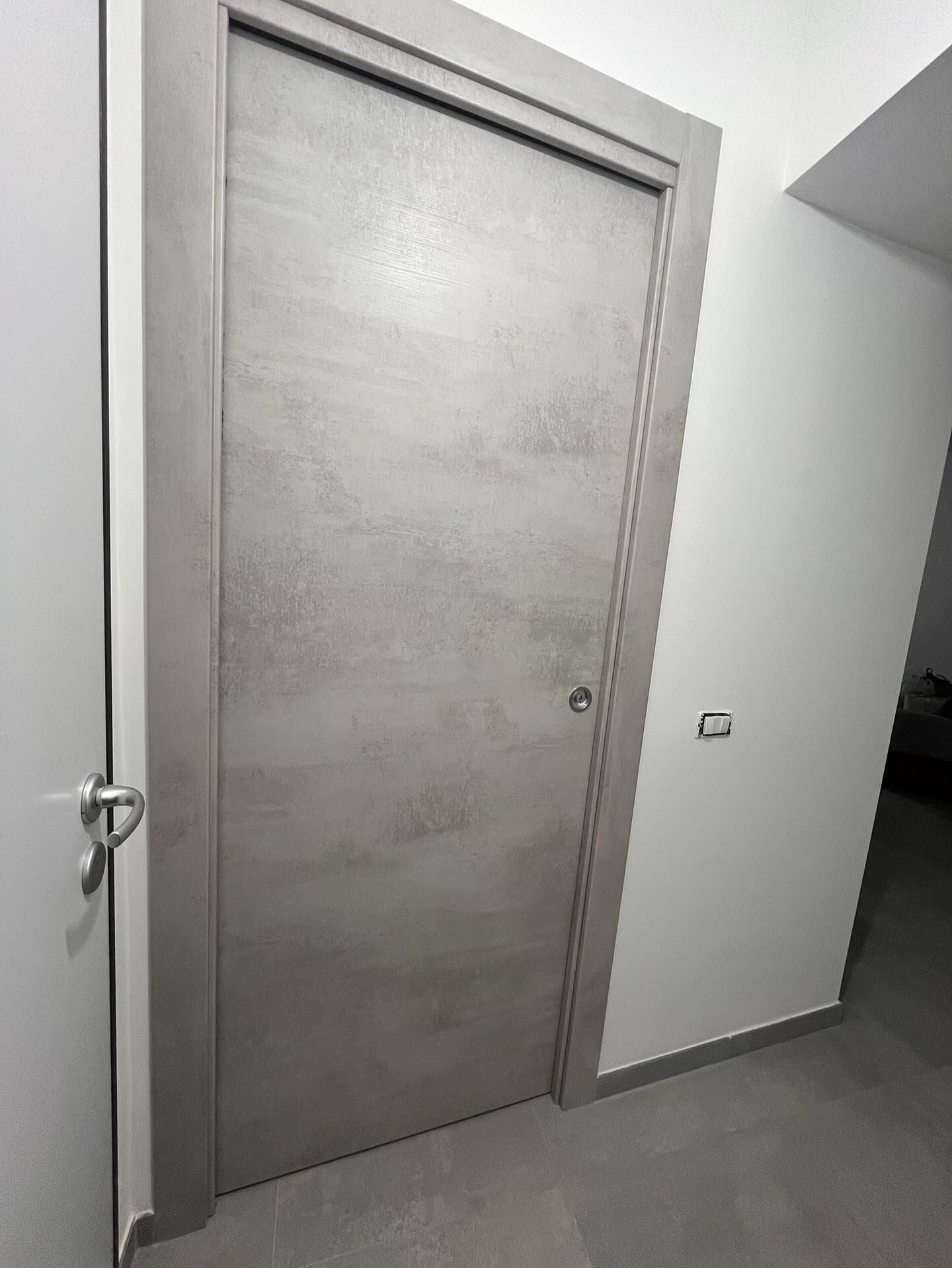 In un corridoio bianco si trova una porta di colore grigio chiaro con una struttura texturizzata e una cornice coordinata, mentre sulla parete adiacente è presente un interruttore della luce.
