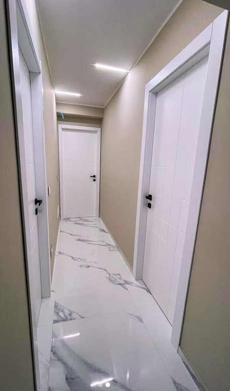 Un corridoio stretto con porte bianche, pareti beige chiaro e pavimento in piastrelle bianche effetto marmo, illuminato da LED a soffitto.