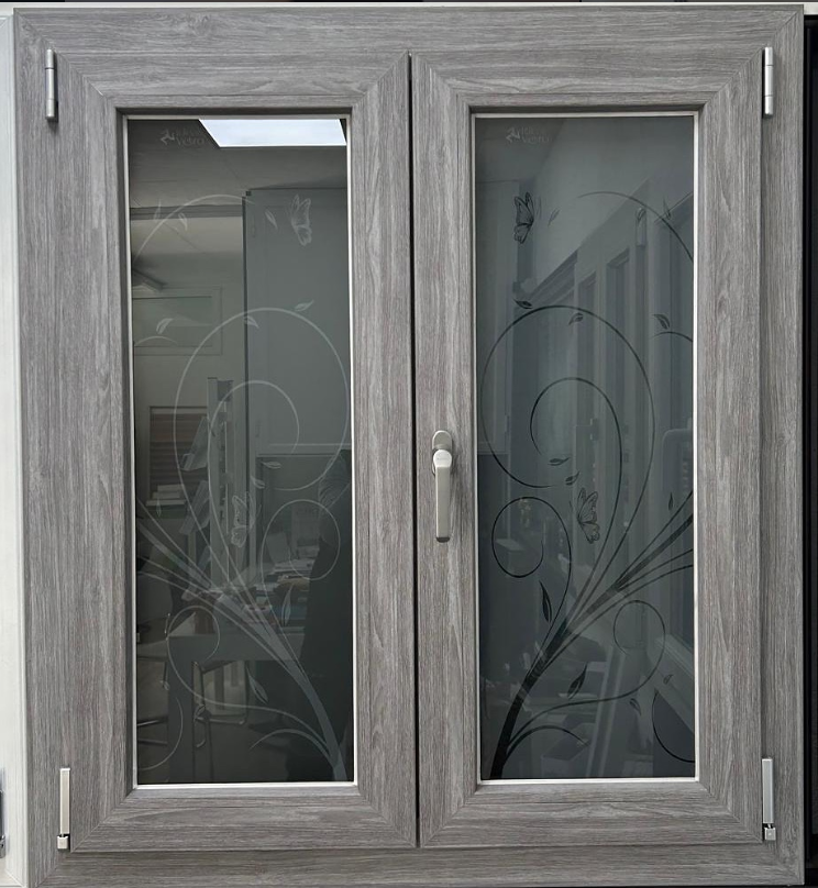 Una finestra a doppia anta con finitura grigia effetto legno e vetro satinato decorativo con motivi floreali scuri e sinuosi.