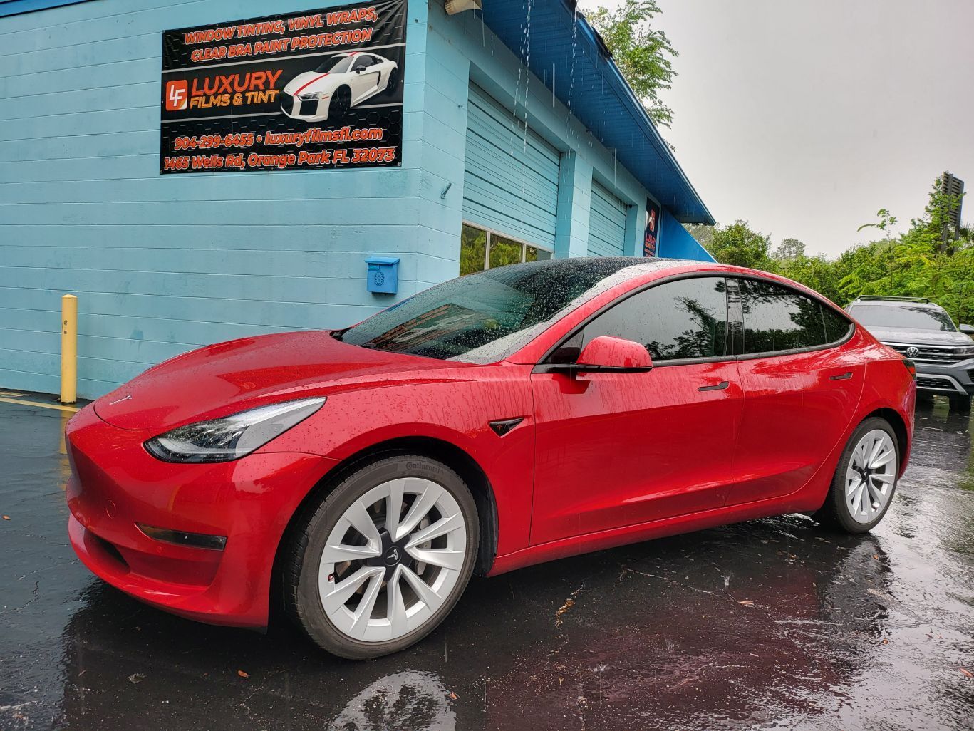 A red tesla model 3 is parked in front of a blue building.
