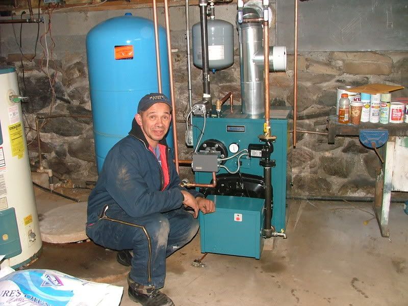 Man in blue jumpsuit kneels beside a teal boiler in a basement. He smiles, with a blue expansion tank behind him.