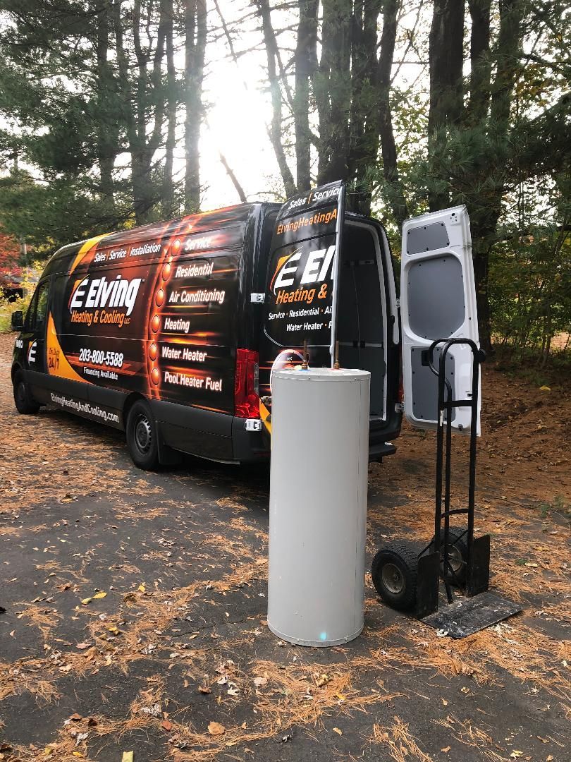 A water heater stands next to a company van with its back door open. A hand truck is nearby.