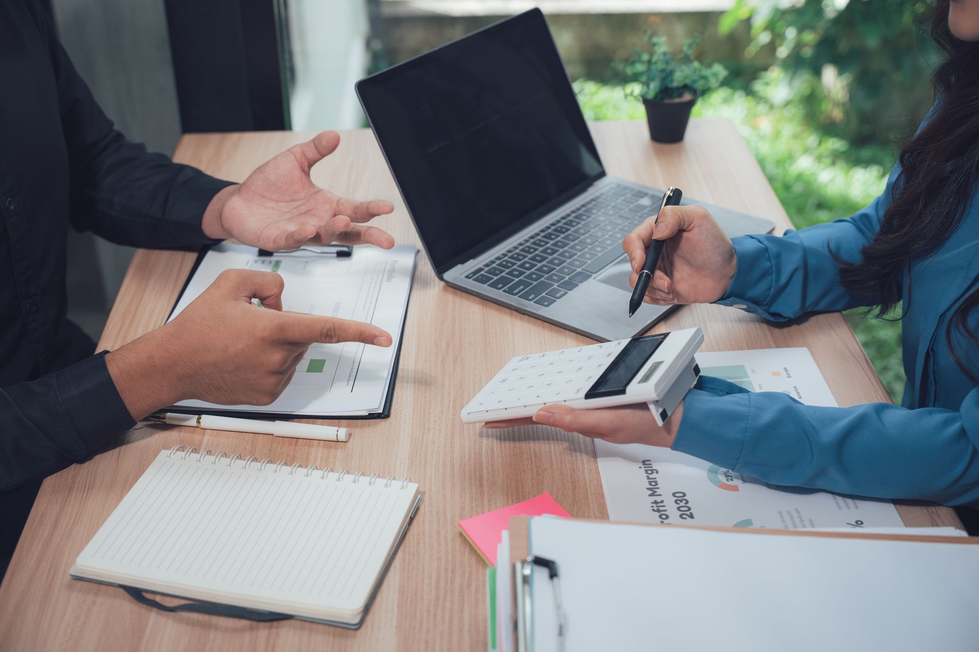 Two people review documents, use calculator and laptop, at a wooden table. Two people review documents, use calculator and laptop, at a wooden table.