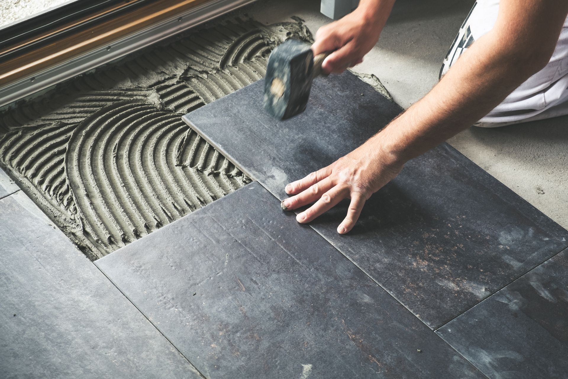 Person using a hammer to set a dark gray tile into wet mortar on a floor.