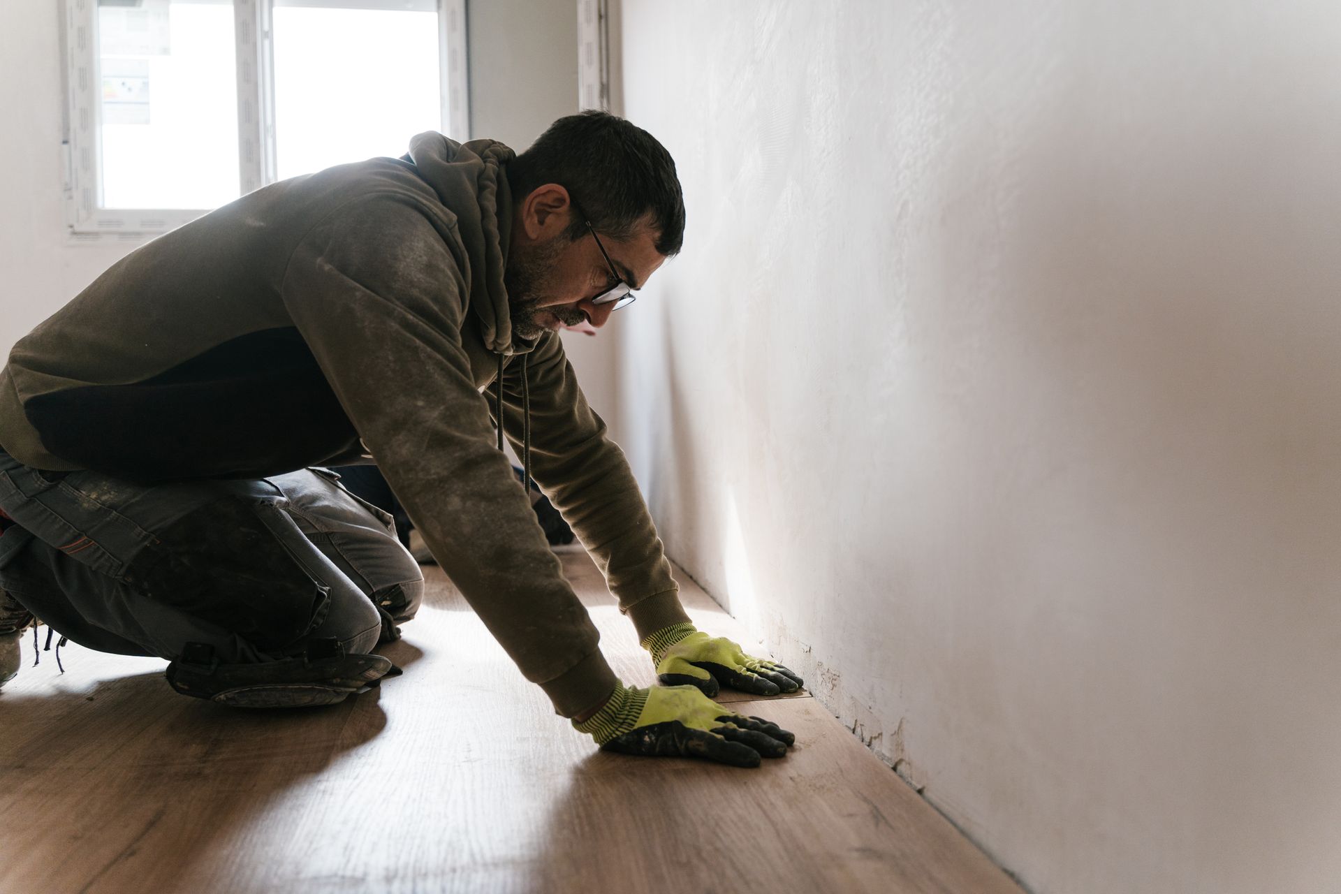 Person on knees installing flooring, near a white wall and window. He wears gloves, hoodie and jeans.