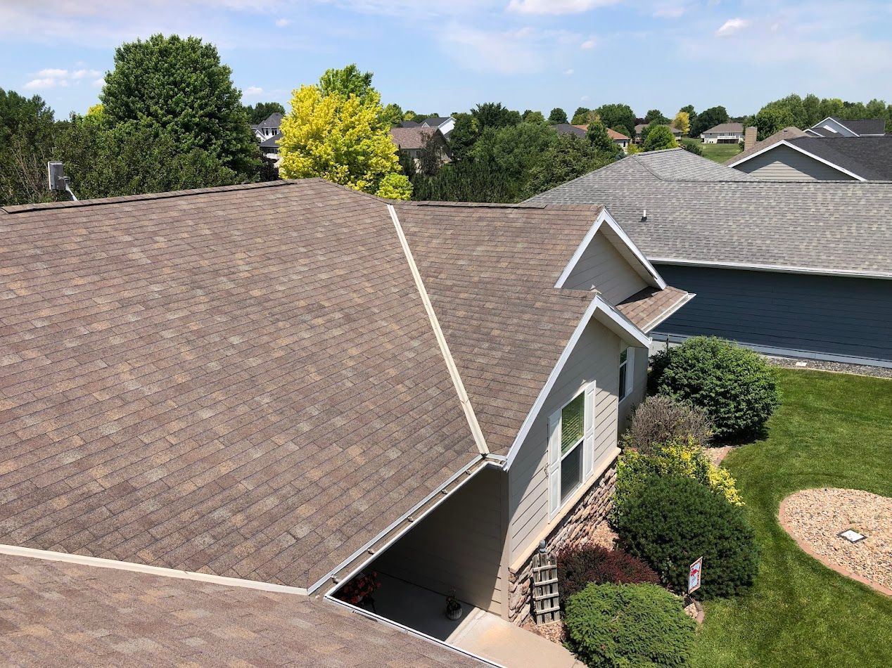 Brown shingle roof of a house with trees and green grass on a sunny day.