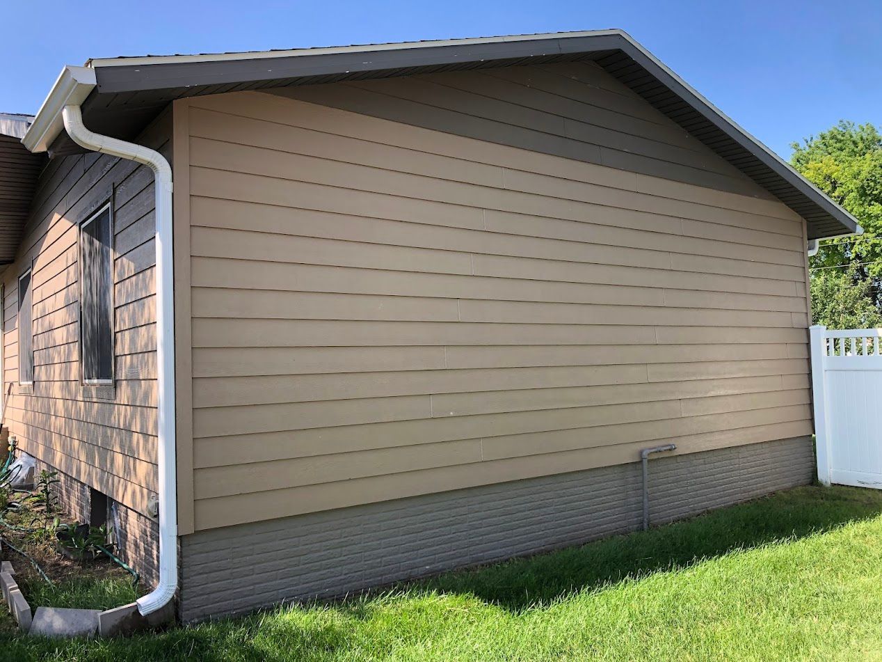 Tan and brown house siding with white gutter and downspout, surrounded by green grass.