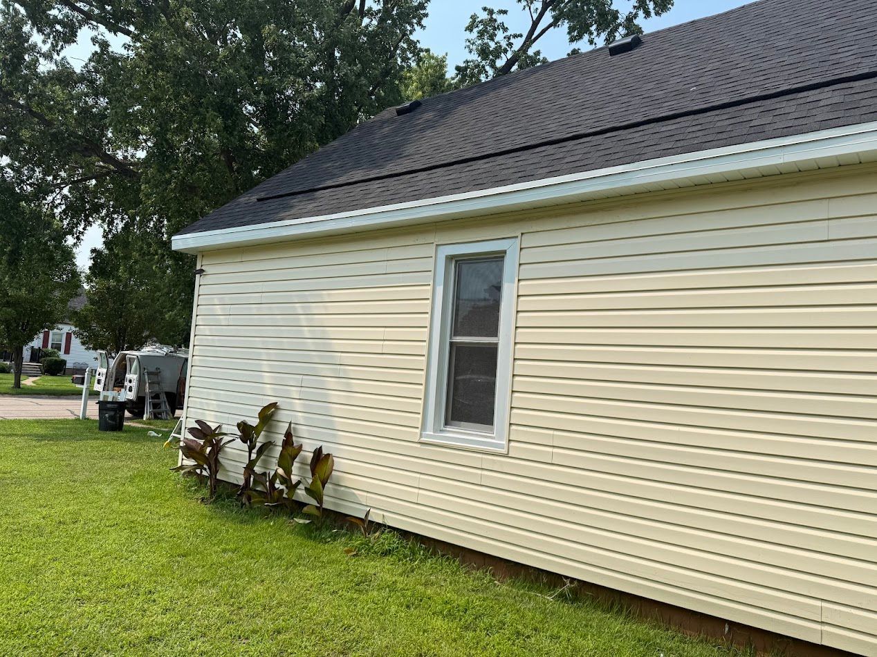 Side of a light yellow house with dark roof and white trim, window, and small plants in front.