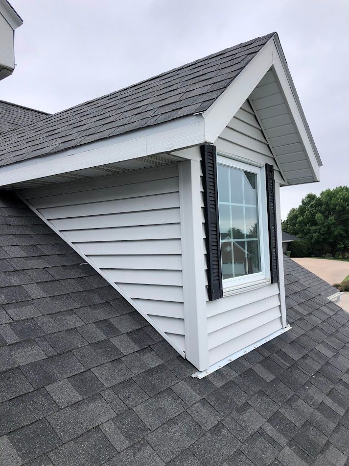 White dormer with window on gray shingled roof, black shutters, white siding and trim.