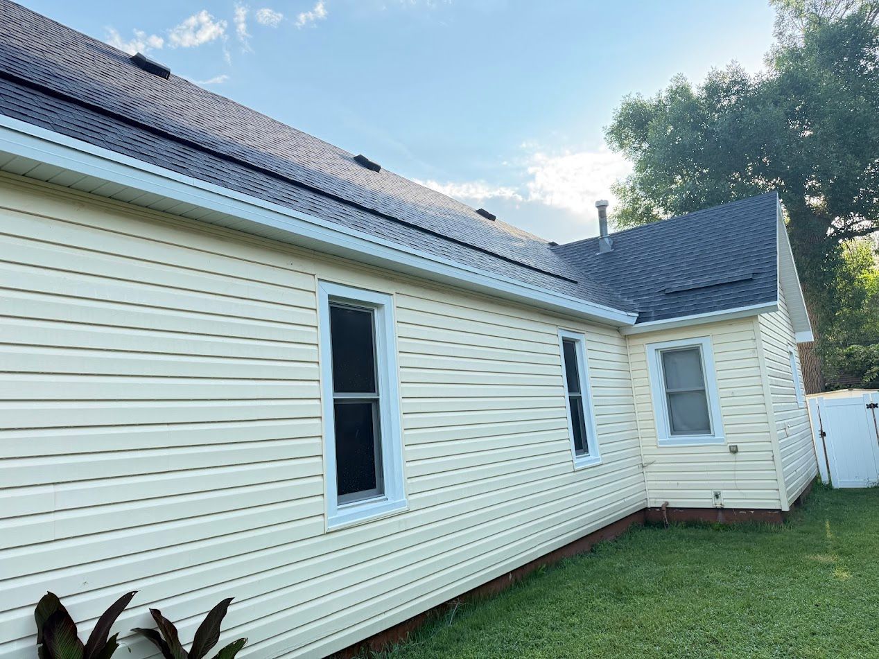 Beige house with a black roof and white trim, with three windows, in a grassy yard.