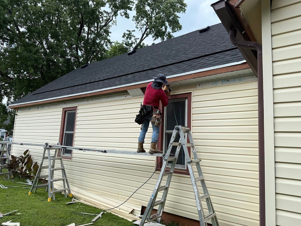 Person installing siding on a house, using a ladder. Beige siding, dark roof, cloudy sky.