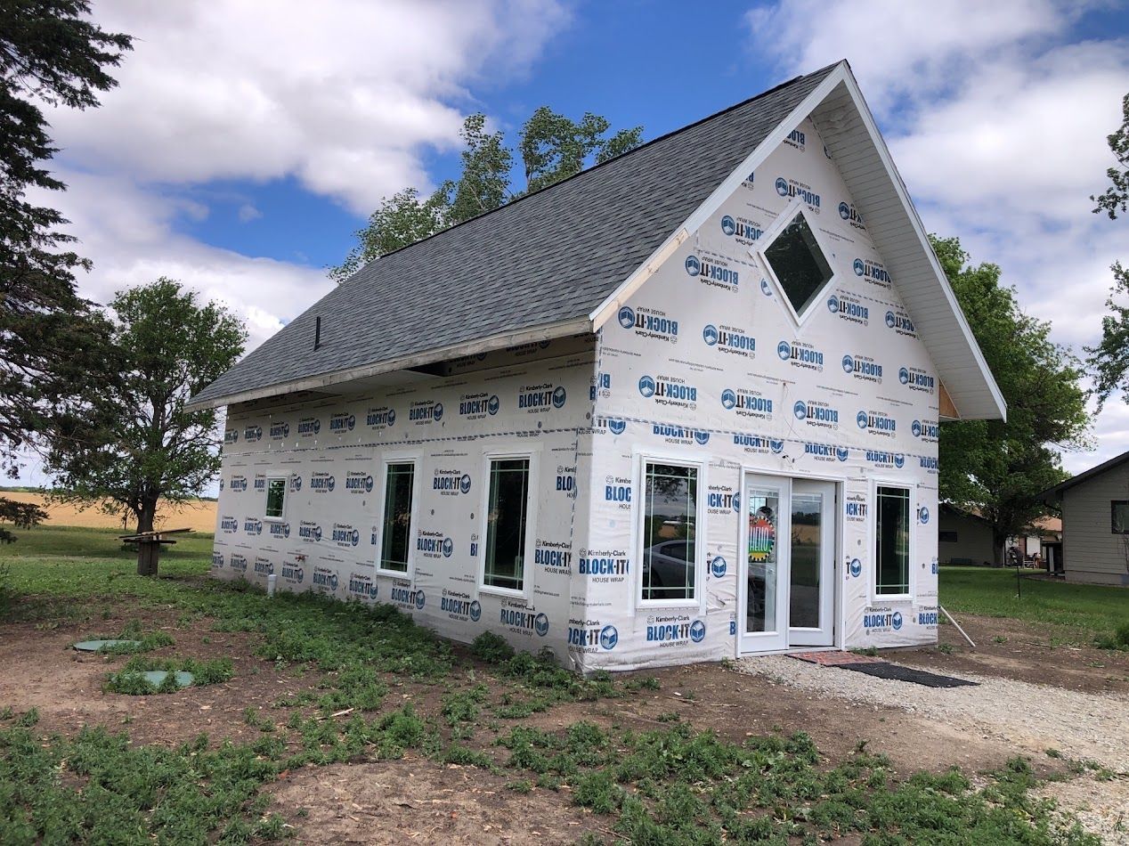 Small, unfinished building with a gray roof and white weather-resistant wrap under a cloudy sky.