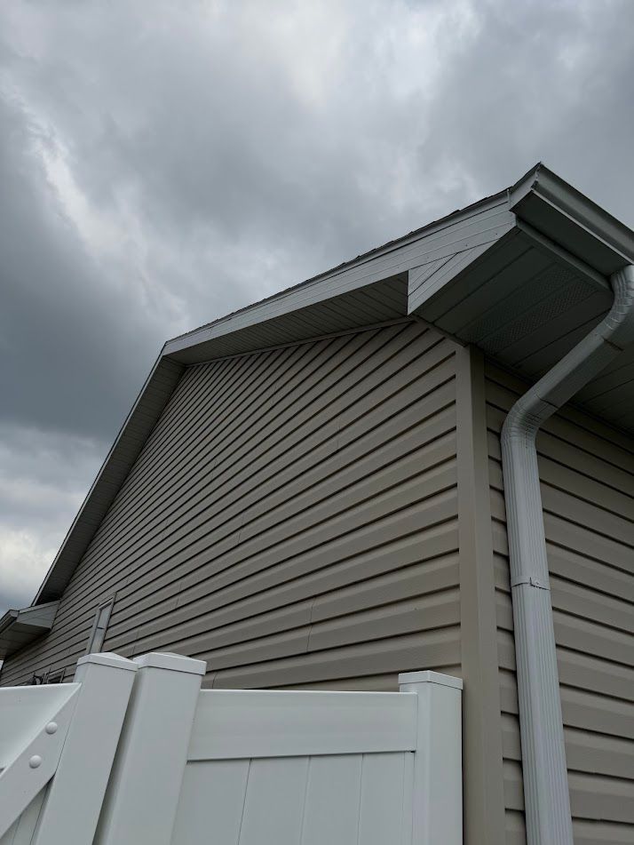 Tan vinyl siding on a building with white gutters and a white fence against a cloudy sky.