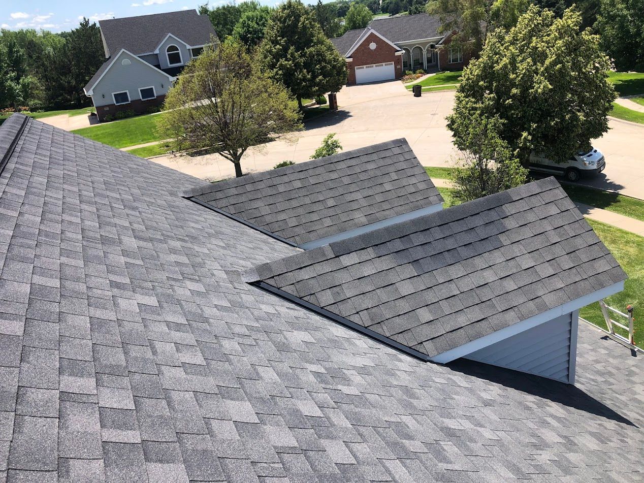 Overhead view of a house roof with gray shingles. Green trees and other houses are in the background.