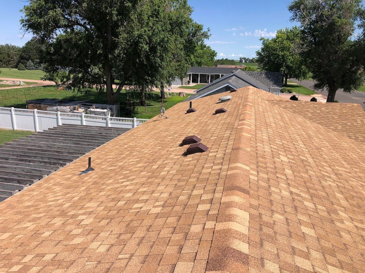 Brown shingle roof with vents against a blue sky, trees, and white fence in background.