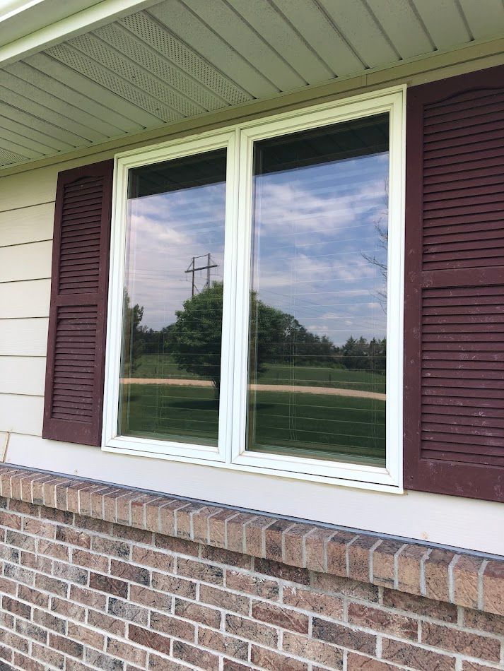 Two-pane window with maroon shutters on a brick and tan siding exterior, reflecting a tree and sky.