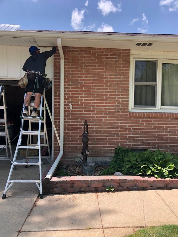 A person on a ladder working on a white gutter on a brick house. Sunny day.