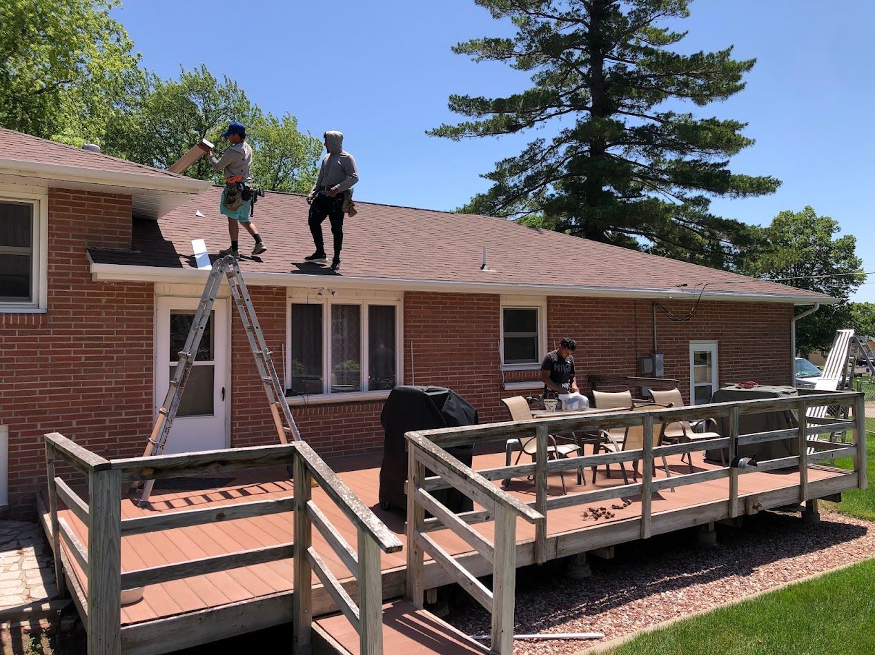 Two workers on a roof, one on a ladder, another on the ground near a deck. Clear, sunny day.
