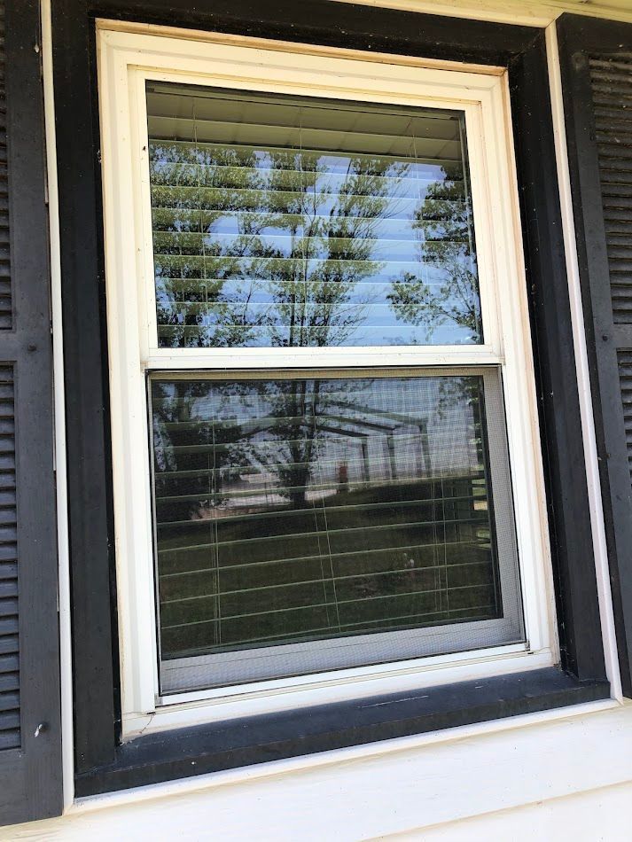 White-framed window with black shutters reflecting trees, sky, and a building; surrounded by white siding.