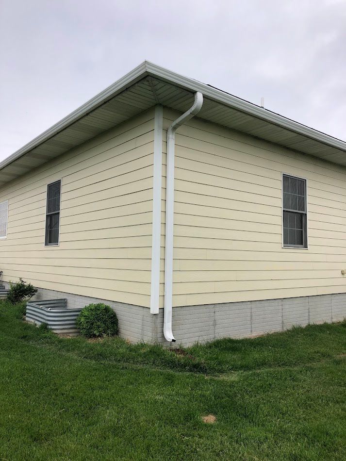 Beige house corner with white gutters, two windows, and green grass. Cloudy sky.
