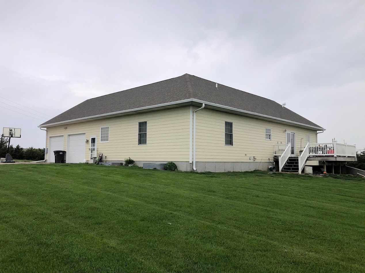 Tan house with a dark roof, white trim, two-car garage, and a deck, on a grassy hill under a cloudy sky.