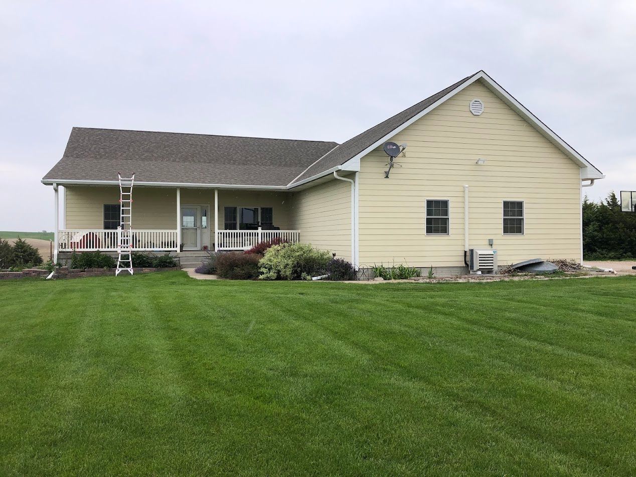 Yellow house with a porch, green lawn, and cloudy sky. A ladder leans against the house.