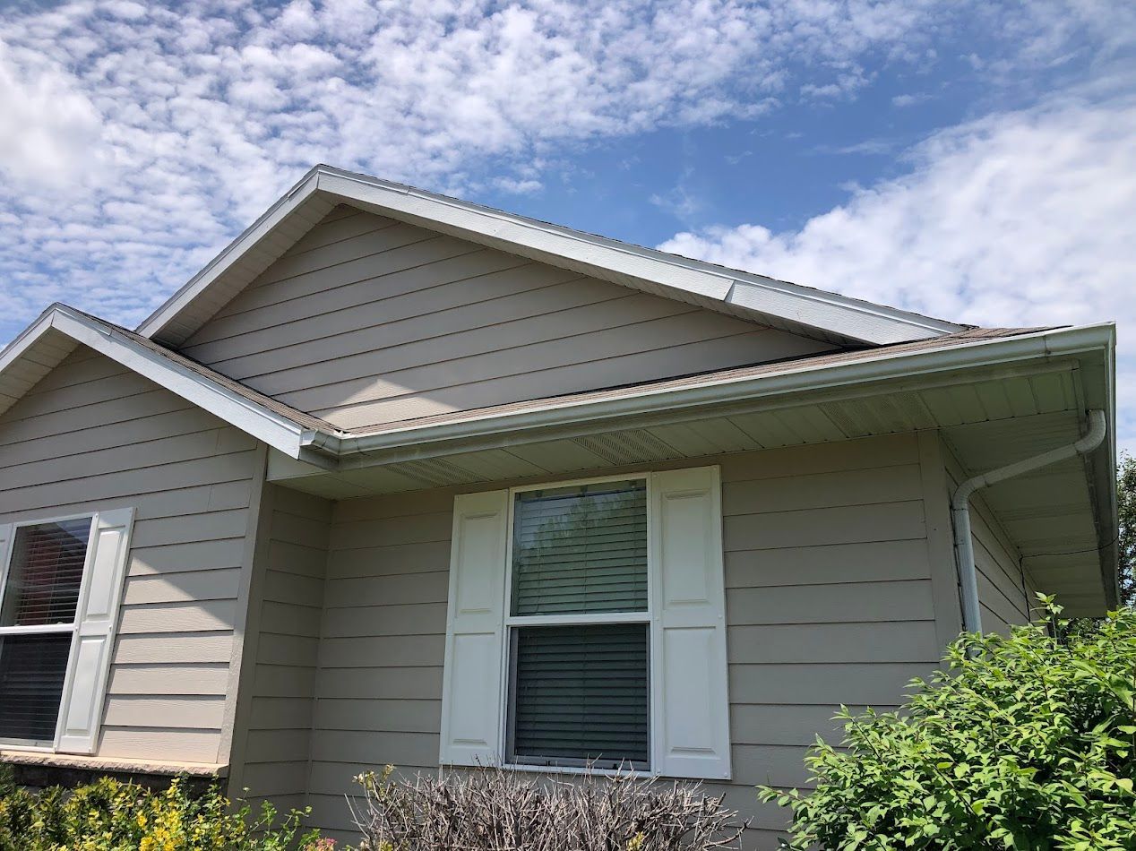 Beige house with white trim, gutters, and shutters against a blue sky with fluffy clouds.