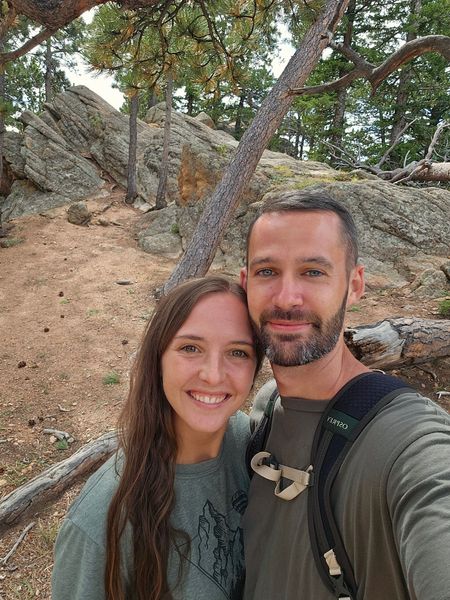 Smiling couple, outdoors, brown-haired woman, man with a beard, forest background, sunny day.