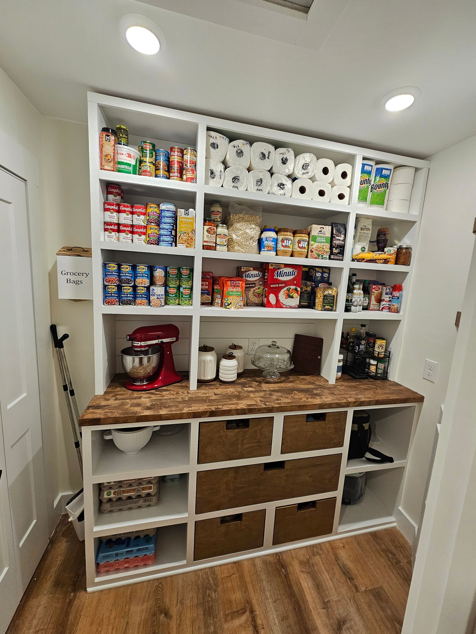 White pantry with wooden countertop, shelves stocked with food and supplies, drawers below.