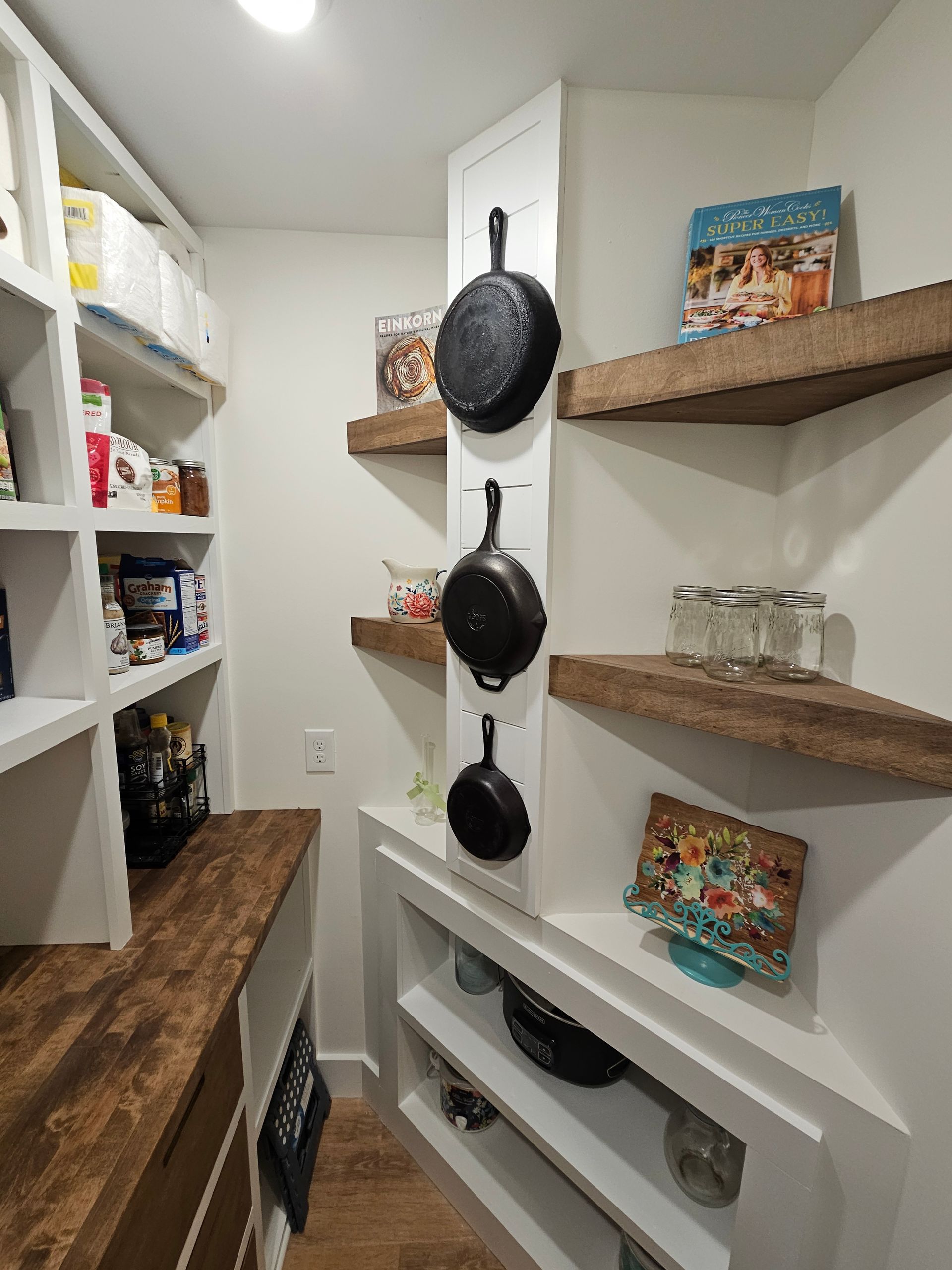 Pantry interior with white shelves and a dark countertop. Cast iron pans hang on a wall.