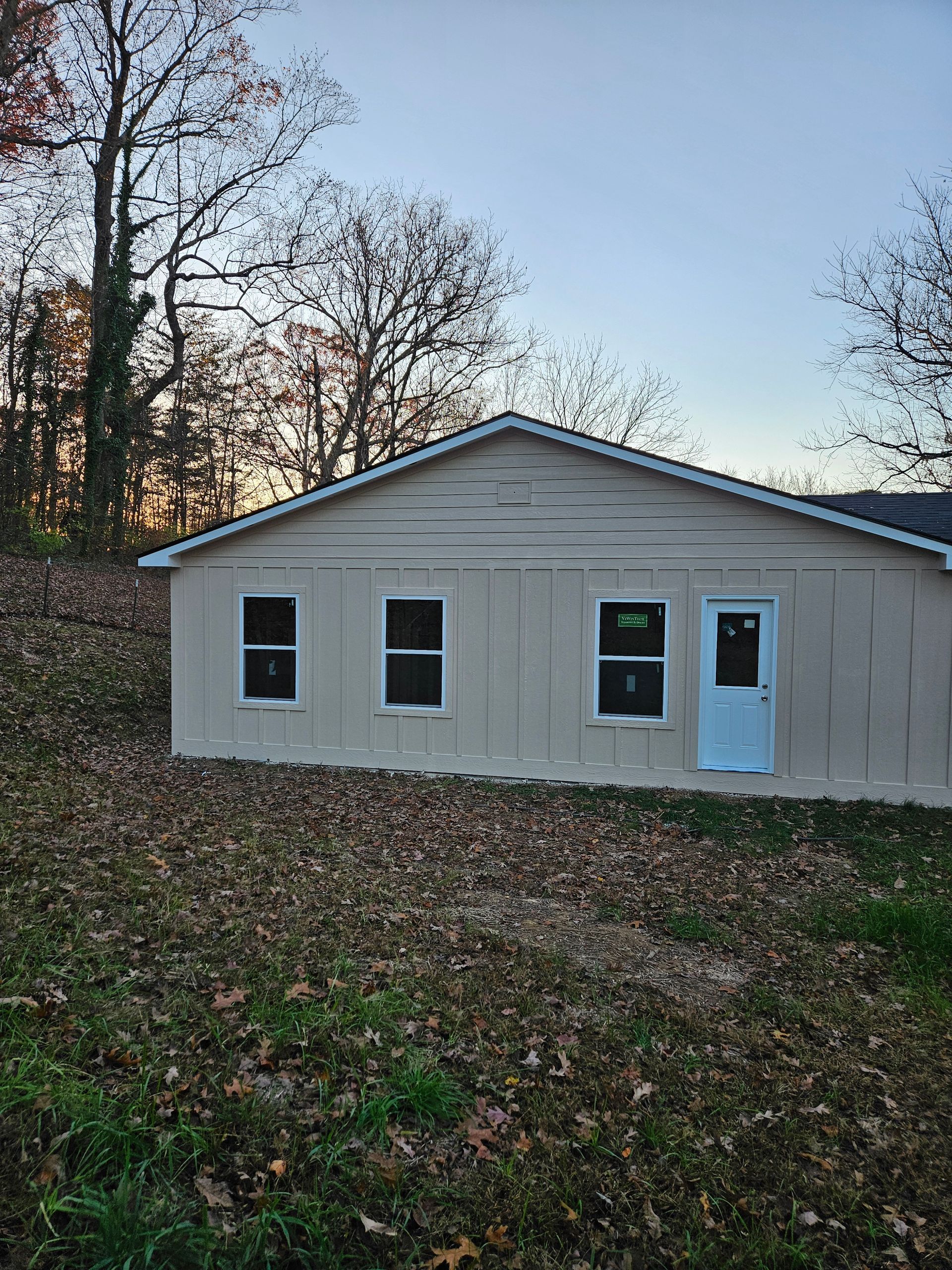 Beige building with white trim, windows, and door, set in a yard covered in fallen leaves, under a dusk sky.