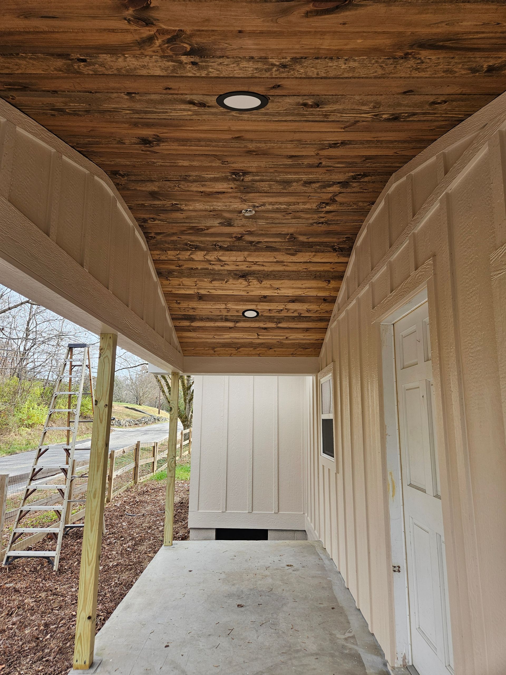 Covered porch with wood ceiling and tan walls, leading to a door and building.
