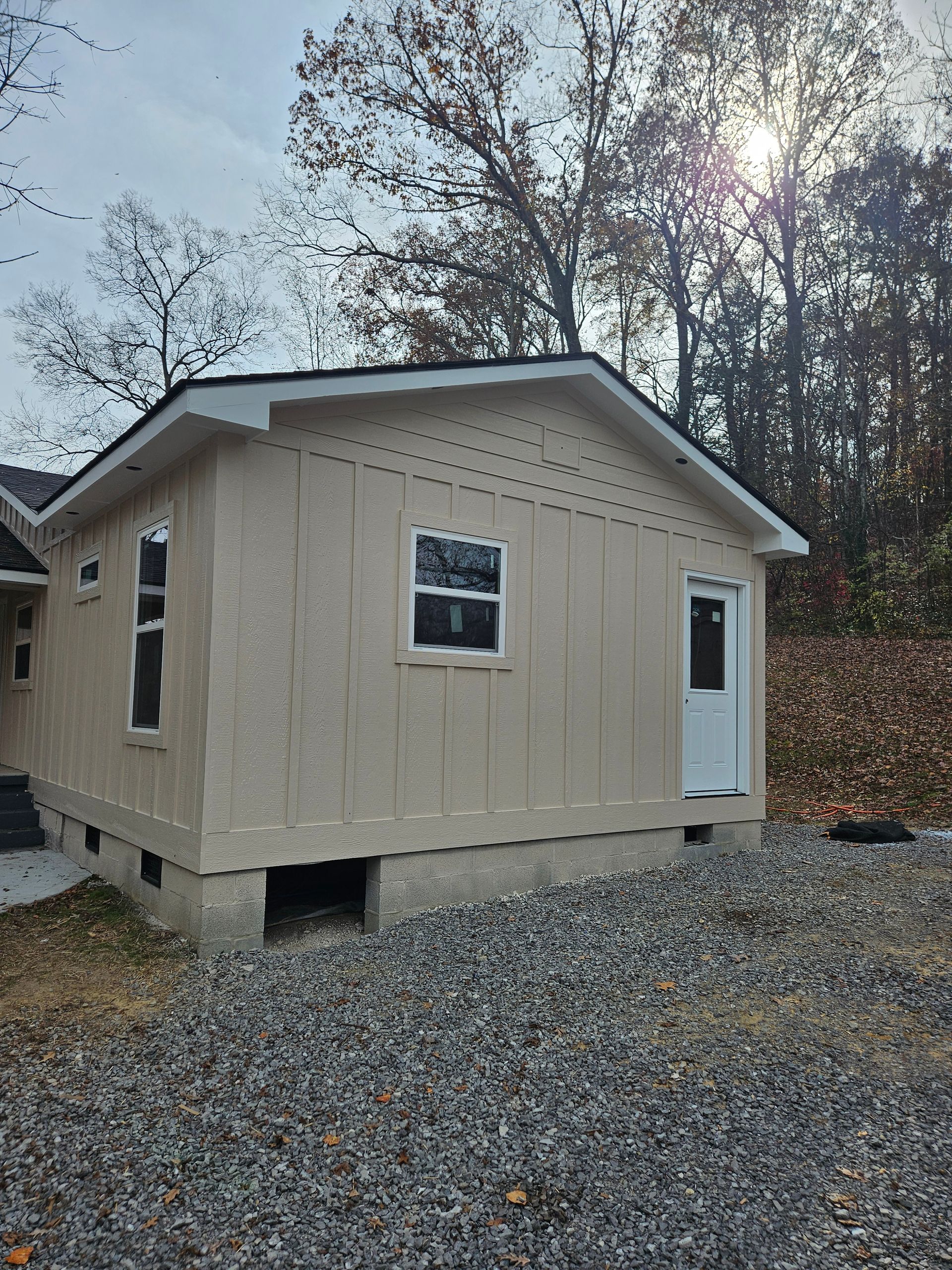 Side of a light tan house with vertical siding, windows, and a white door, set in a gravel yard.