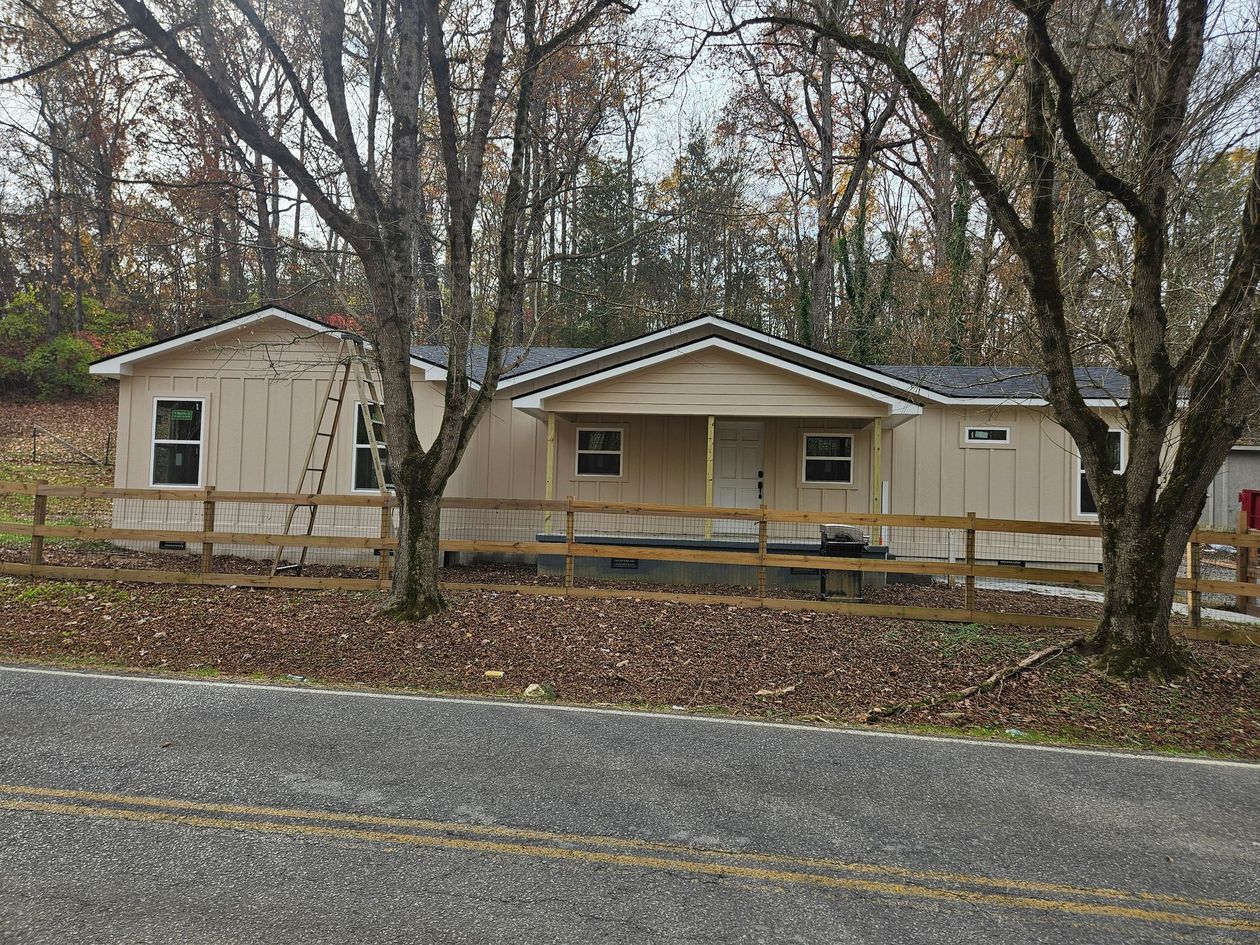 Beige house with a porch and a wooden fence, set amongst trees. A ladder leans against the house.